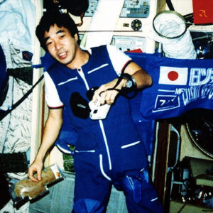 A man in a blue and white coveralls floats inside a space station module surrounded by instruments and stowage bags