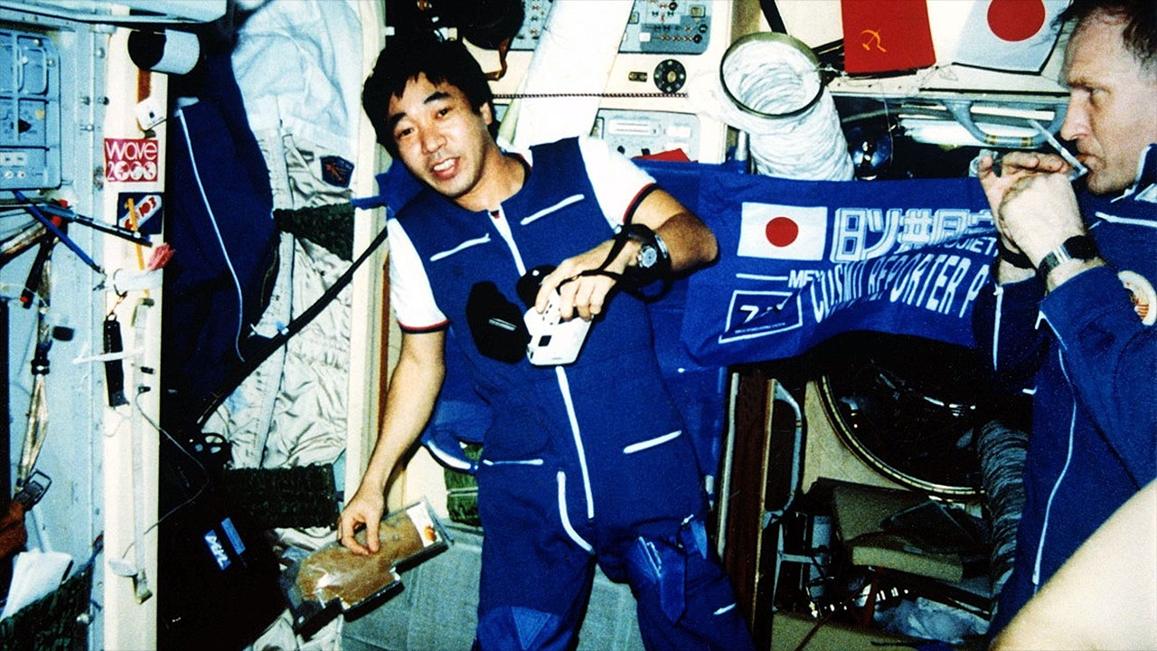 A man in a blue and white coveralls floats inside a space station module surrounded by instruments and stowage bags
