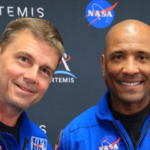 Two men in a blue flight suits pose for a photo in front of a black wall with printed logos.