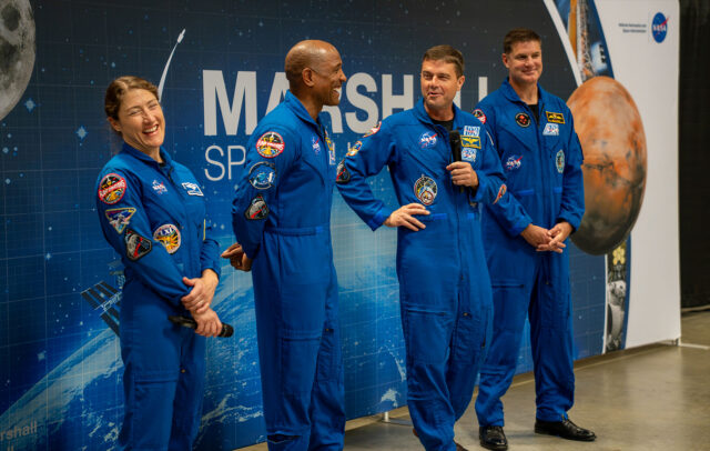 Three men and a woman, all wearing blue flght suits, stand together at the front of a room with a backdrop reading "Marshall Space Flight Center" behind them.