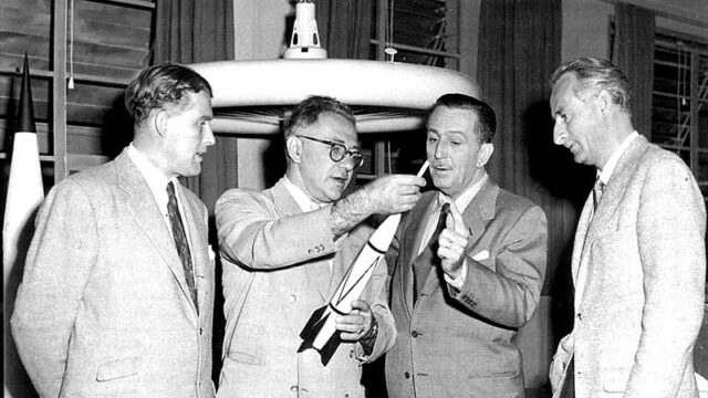 Black and white photo of four men in business suits examining models of future proposed spacecraft.
