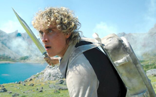 young man with curly blonde hair holding a weapon in an outdoor arena