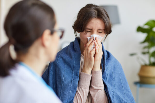 A woman blows her nose into a tissue while a doctor stands before her.