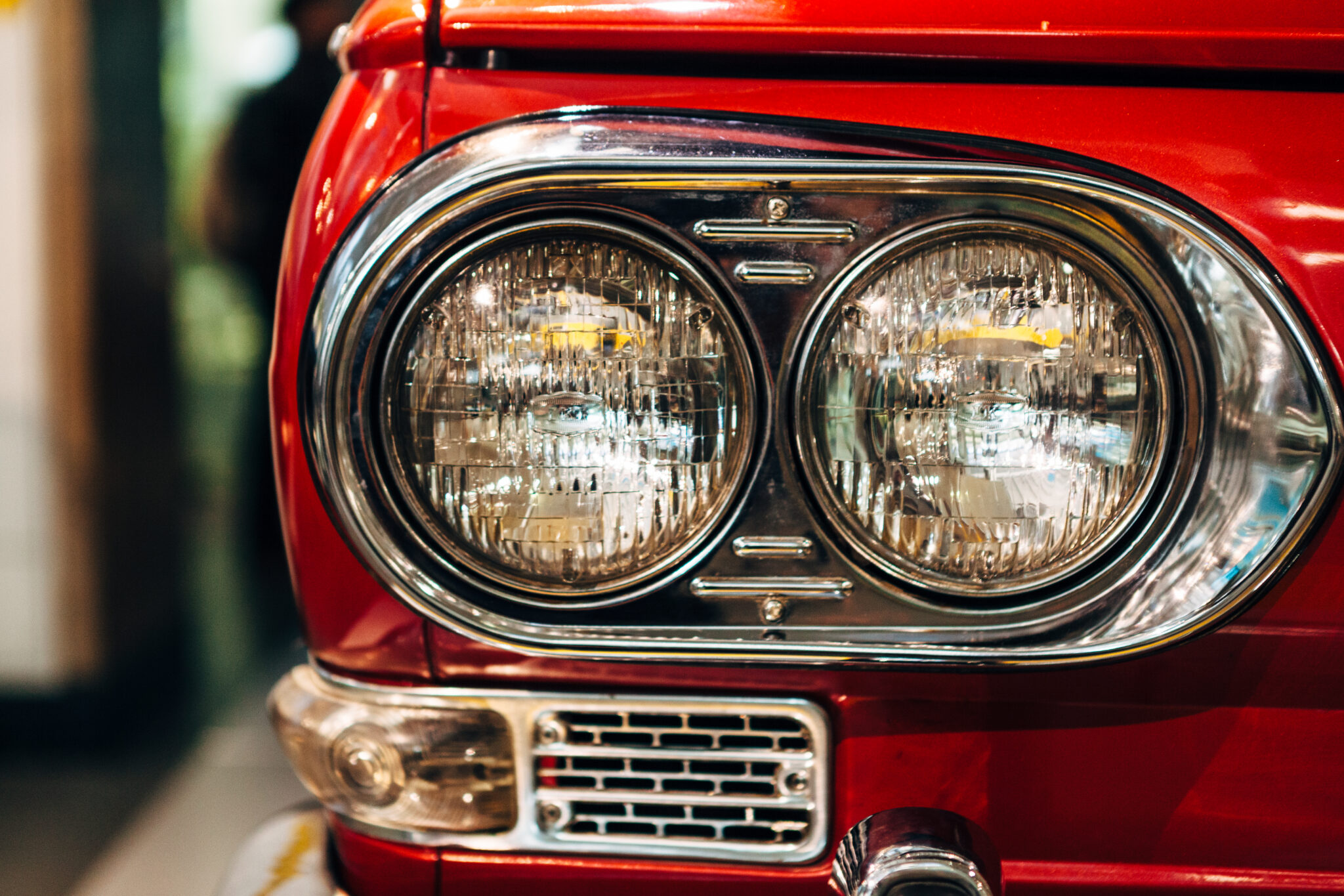 A close up view of the front right side of a vintage red Japanese car featuring twin circular headlights surrounded buy chrome.