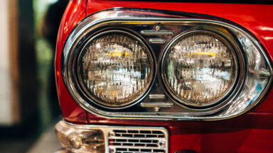A close up view of the front right side of a vintage red Japanese car featuring twin circular headlights surrounded buy chrome.