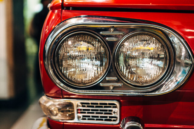 A close up view of the front right side of a vintage red Japanese car featuring twin circular headlights surrounded buy chrome.