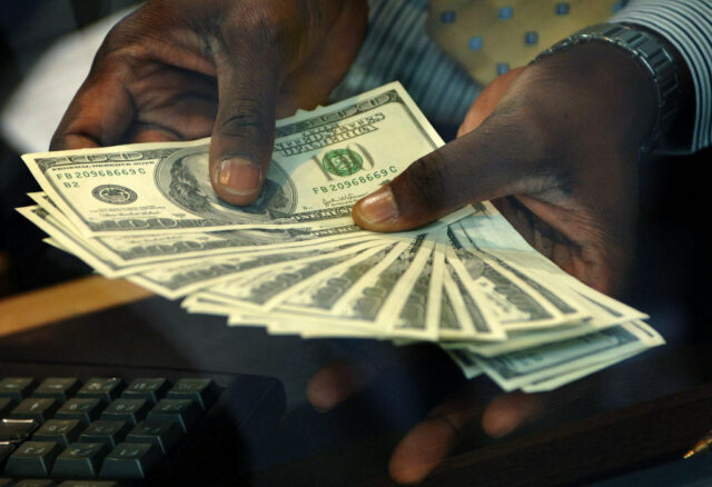 The hands of a man wearing a suit fans out $100 bills near a keyboard.