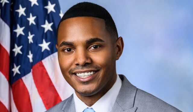 A man smiles in a suit in front of an American flag