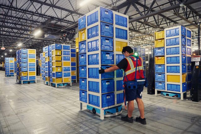 Wheeled mobile robots move tall inventory pods filled with items inside an Amazon fulfillment center. A human employee wearing an orange safety vest is steadying one of the racks.