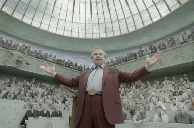 aging man in cowboy suit standing in a large atrium in front of a crowd, arms held wide