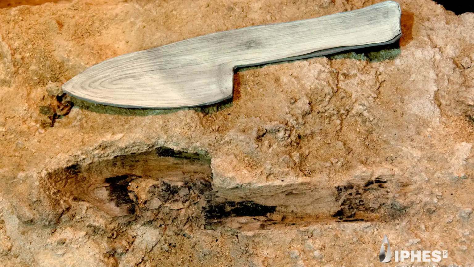 Image of a stone block with the imprint of a device with a blade and handle, with a model of a wooden version of the object that formed it just above.