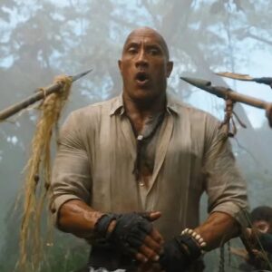 large bald very muscular man in tight shirt standing open mouthed while surrounded by jungle natives brandishing spears