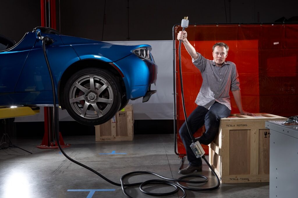young Elon Musk sitting on a wooden crate while brandishing a charger in one raised hand. A blue early Tesla prototype is to the left.
