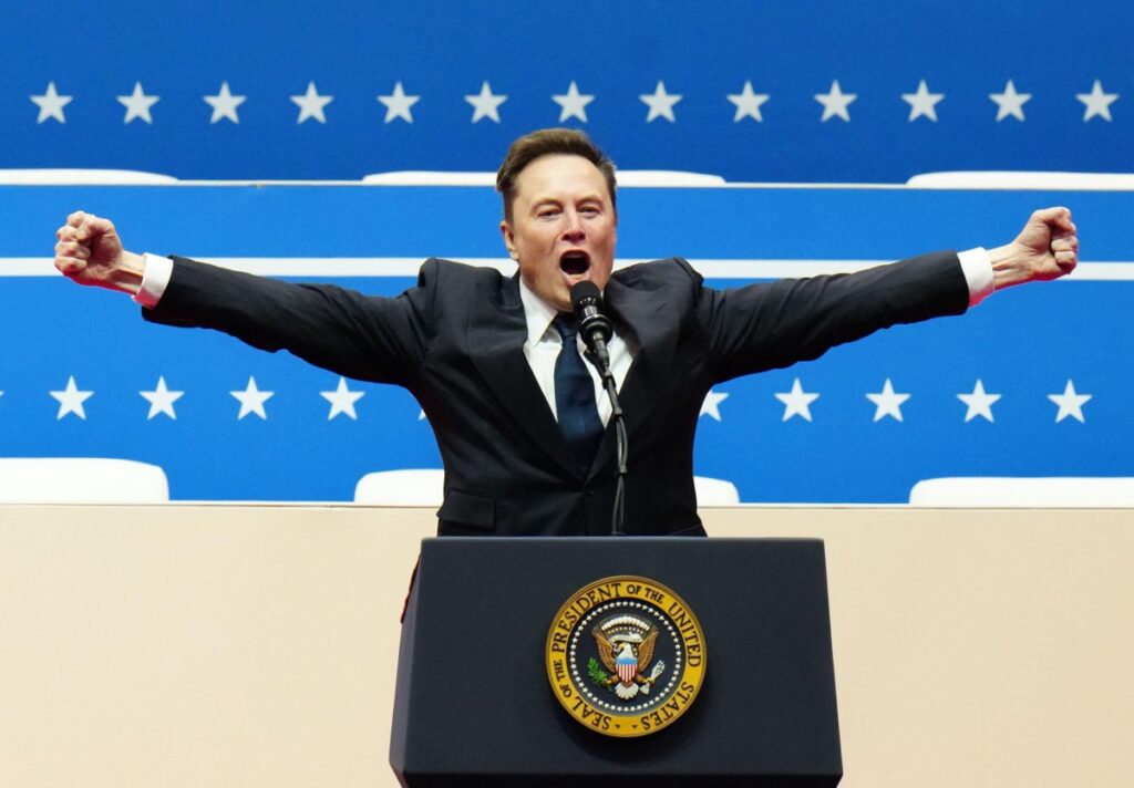 Older Musk in dark blue suit and tie, standing at podium at political event, arms spread wide, as he speaks into a microphone