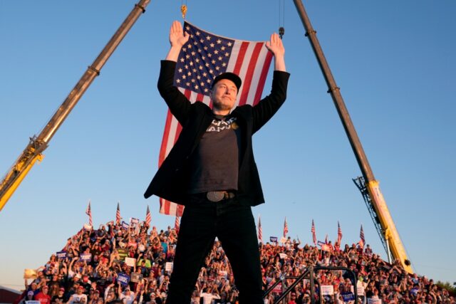 elon must in jeans and t shirt and denim jacket with a black MAGA hat in front of a US flag with arms raised as a crowd behind him cheers