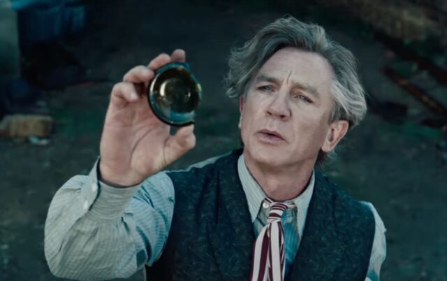 Middle aged man with wild gray hair in 1950s clothes holding up the broken off bottom of a green glass bottle