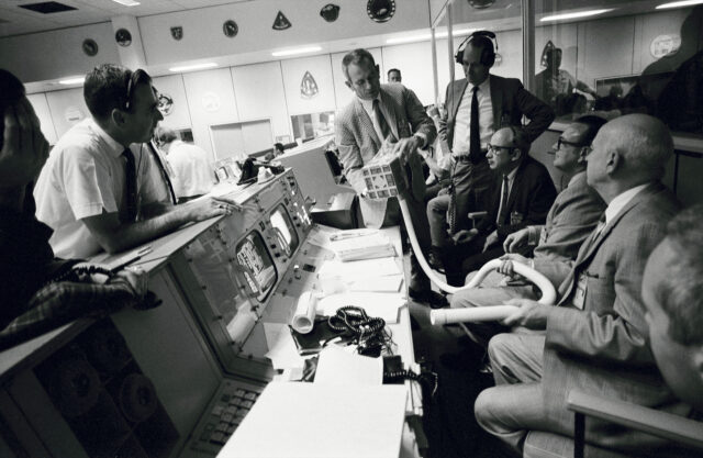 a black and white photo of several men sitting at consoles while inspecting makeshift device with tube extending from one side.