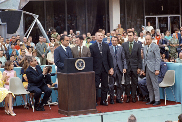 a photo of several men standing on an outdoor stage, surrounded by an audience. One man is standing behind a podium with the seal of the President of the United States.