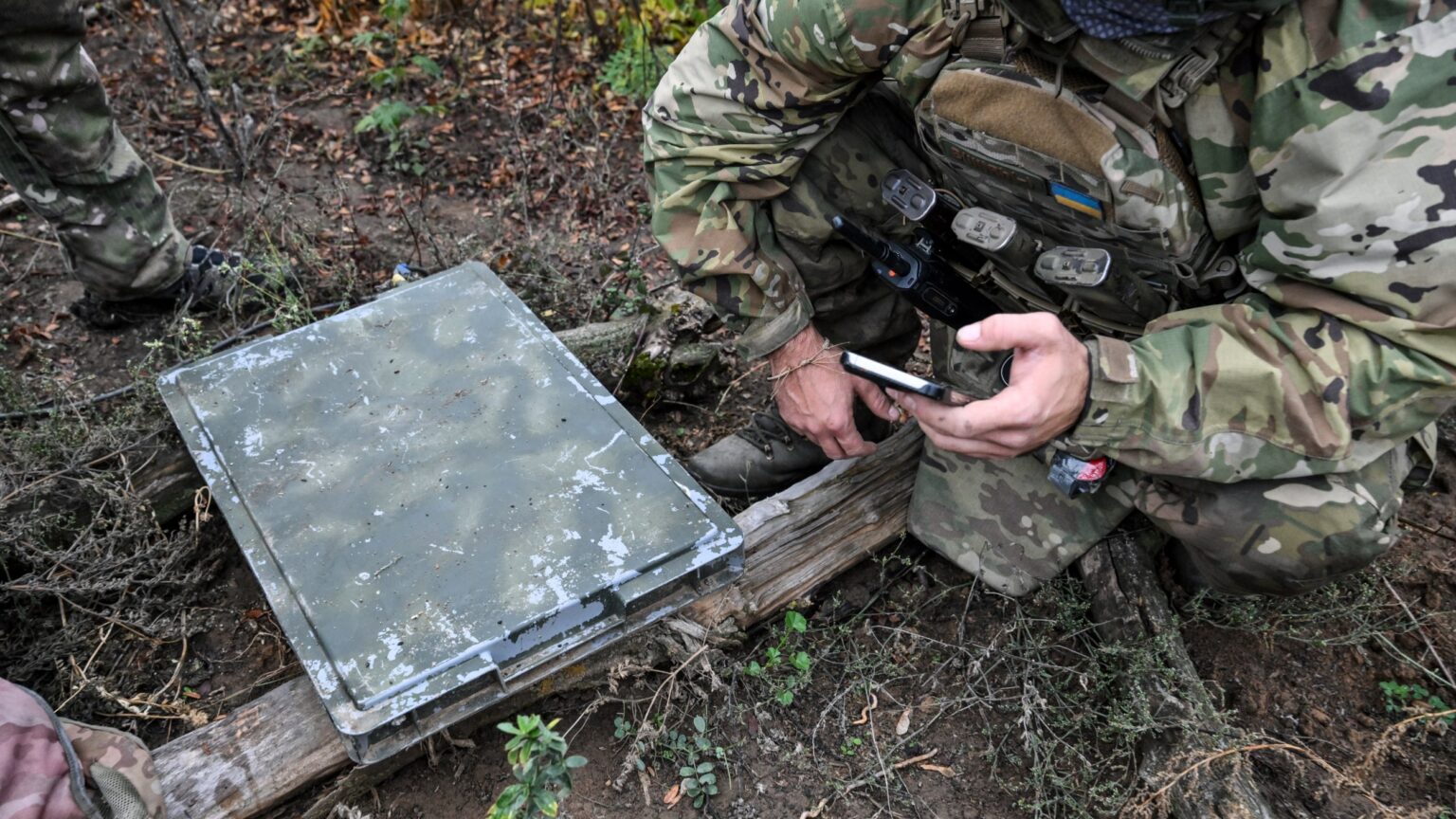 A soldier wearing combat fatigues kneels next to a Starlink terminal while looking at a smartphone.