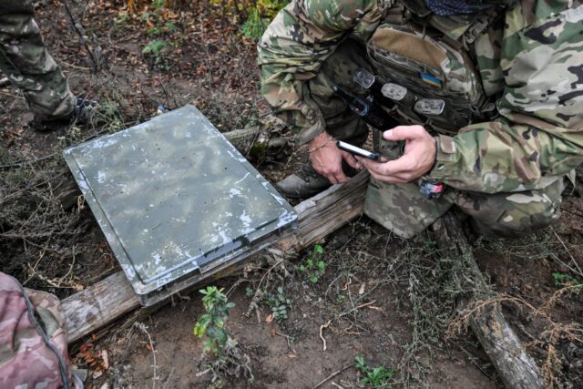 A soldier wearing combat fatigues kneels next to a Starlink terminal while looking at a smartphone.